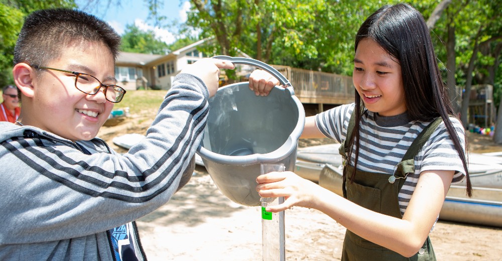 Two children testing water turbidity, with them holding a bucket and pouring water into a tube.