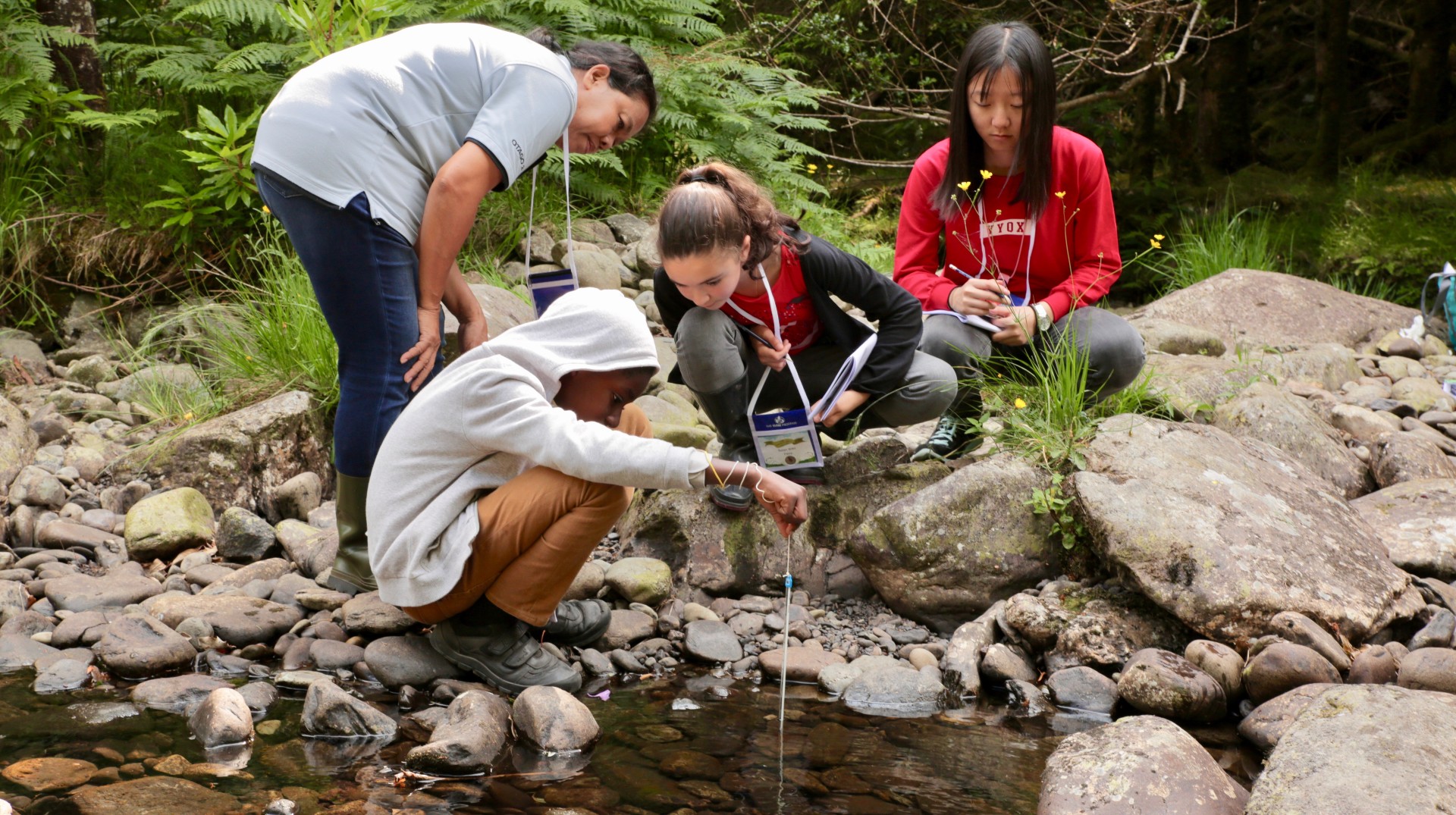 Four people crouch down near a river. One of the people collects a water sample with a glass or plastic pipette.