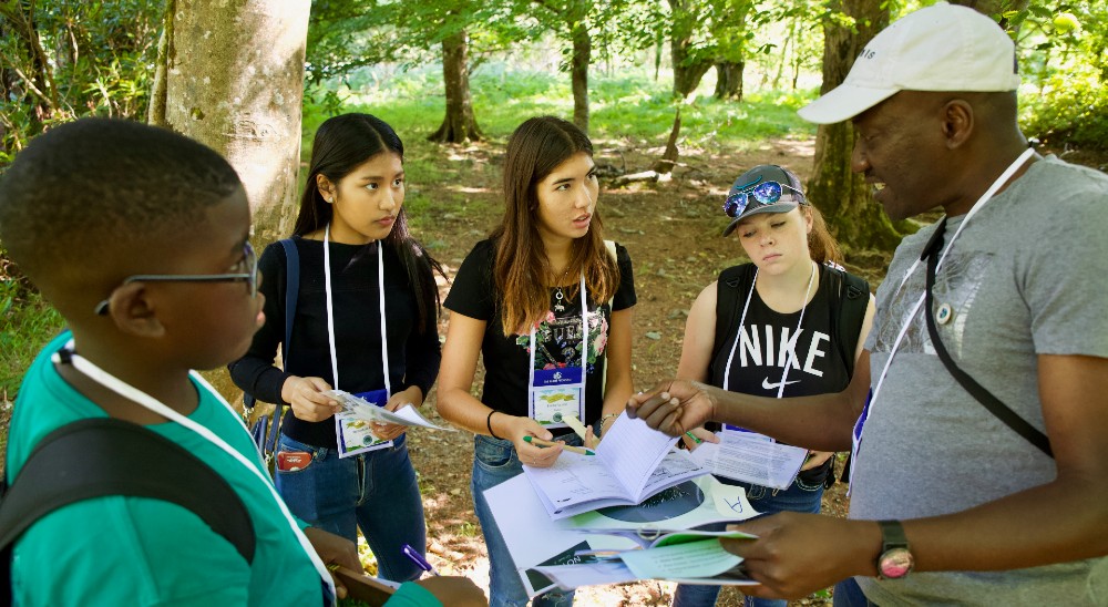 A scientist explains a lesson to several students outside under some trees.