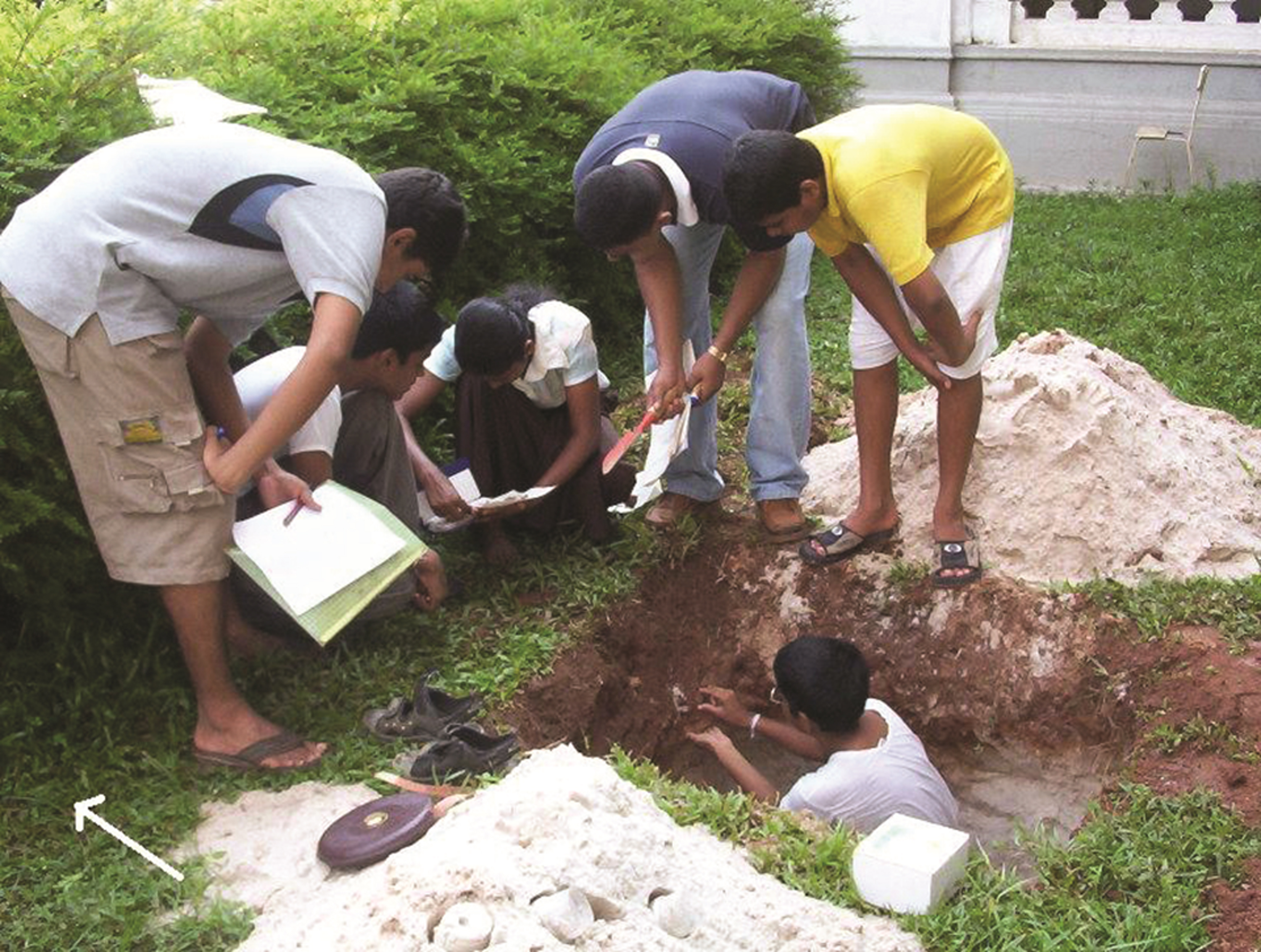 Students standing around a hole, holding science equipment and papers, as one student digs in the hole.