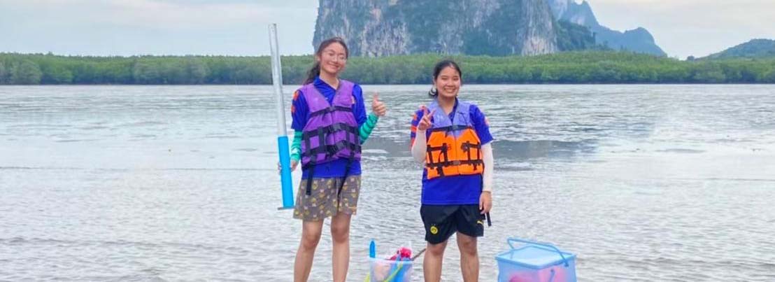 Two students wearing life jackets stand by a lake, holding a tube for measuring water data.