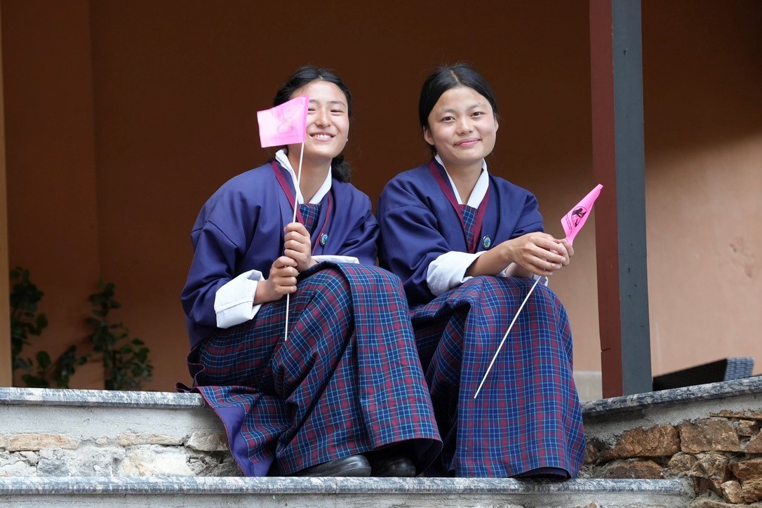 Two women sit on a concrete stoop. They are both smiling and holding pink flags.
