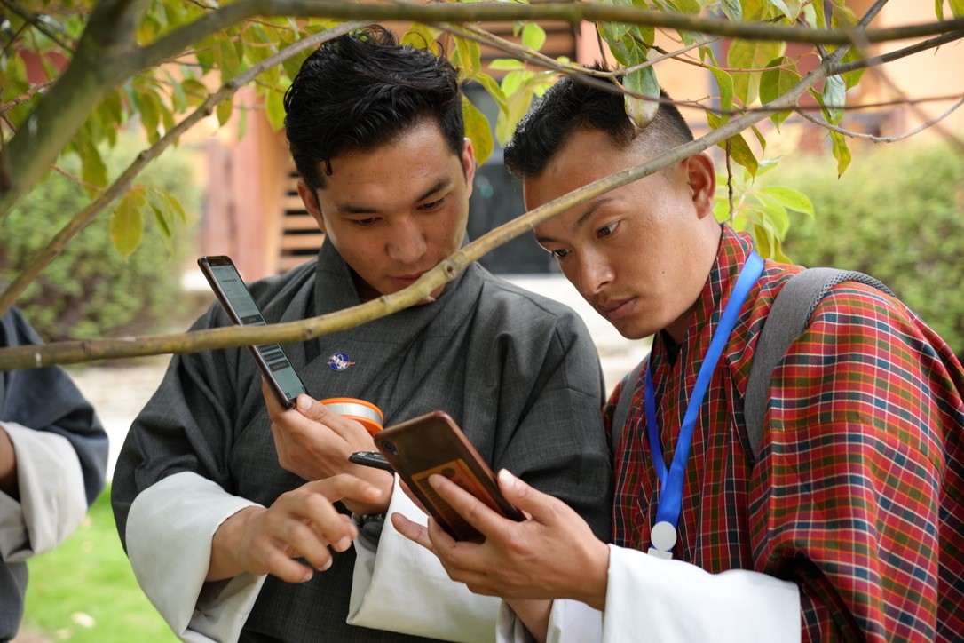 Two men stand under a tree outside. Each of the men hold phones and both men are staring at the phone of the man on the right. 