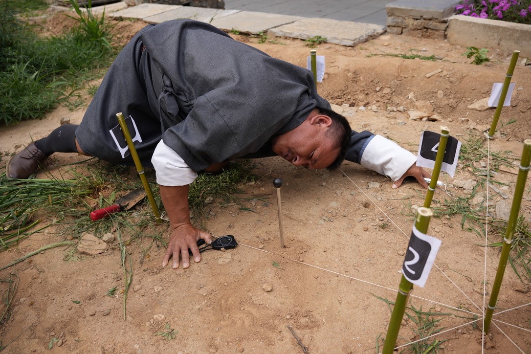 A man kneels in dirt beside several wooden sticks placed in the ground. Each stick is numbered.