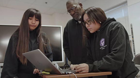 Two students and a teacher look at a computer screen.