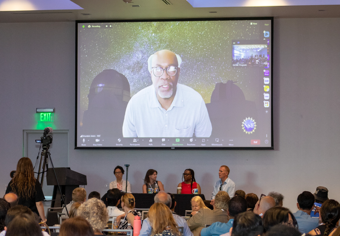 A crowd of people watch a NASA scientist deliver a speech on a large screen.