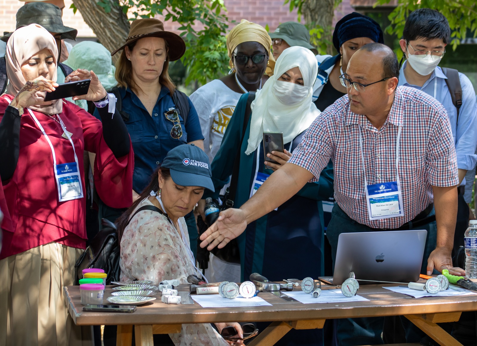 A group of teachers stand around a table covered with instruments, while a presenter speaks to a group.