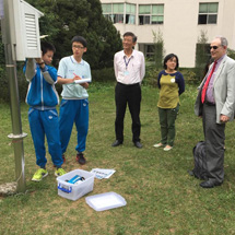 A young boy student demonstrates a weather station for several adults outdoors.