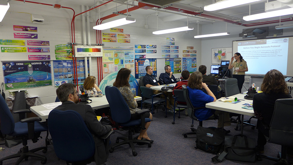 A group of people watch a GLOBE trainer presenting in a classroom.