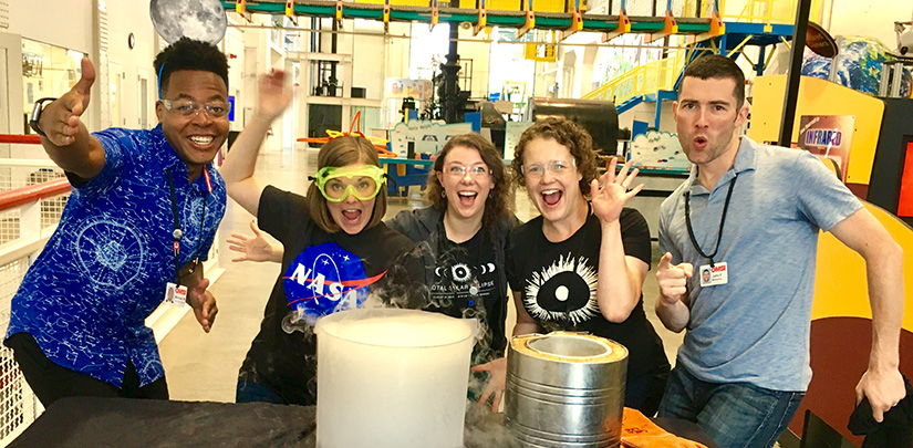 Four students and an adult smile around a table in front of a science experiment, while wearing goggles.