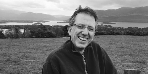 Man smiling with a lake in the background in black and white. 