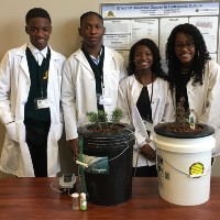 Four students are smiling and dressed in white lab coats. There is a table in front of them that has two buckets, both filled with soil and plants. Behind the students is a poster.