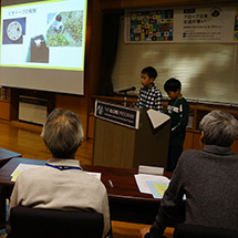 Two boys stand behind a podium and give a PowerPoint presentation to a group.