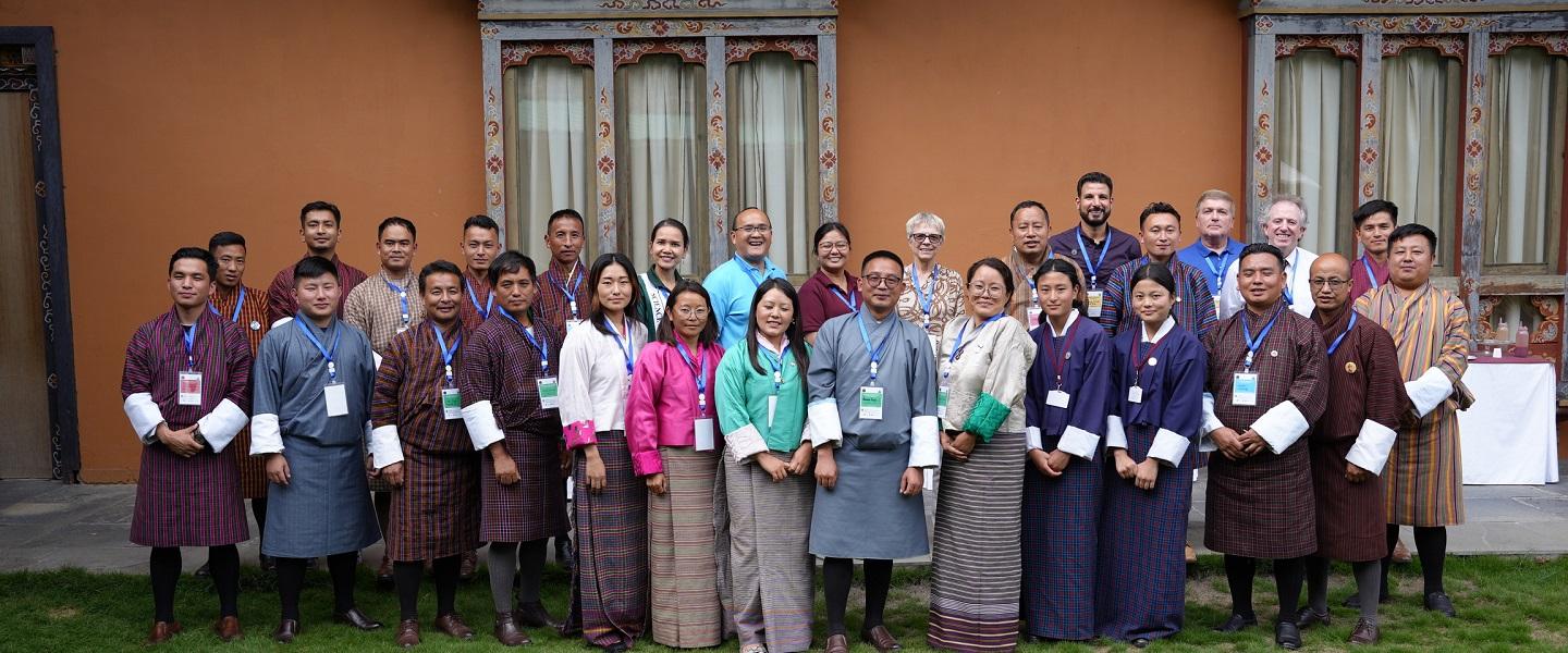 A group of people stand and smile for a photo out of doors. A building is their backdrop and green grass is in the forground.