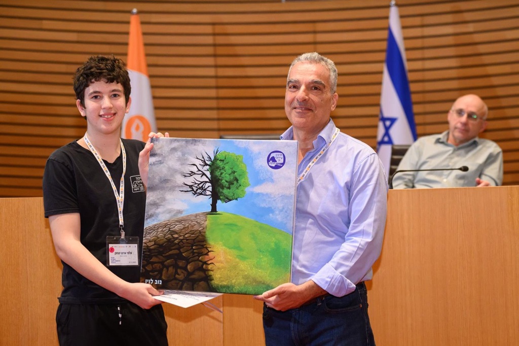 Two people holding a painting of a tree. Half of the painting is of a dead tree, infertile ground, and polluted skies. The other half has leaves, grass, and clear skies.