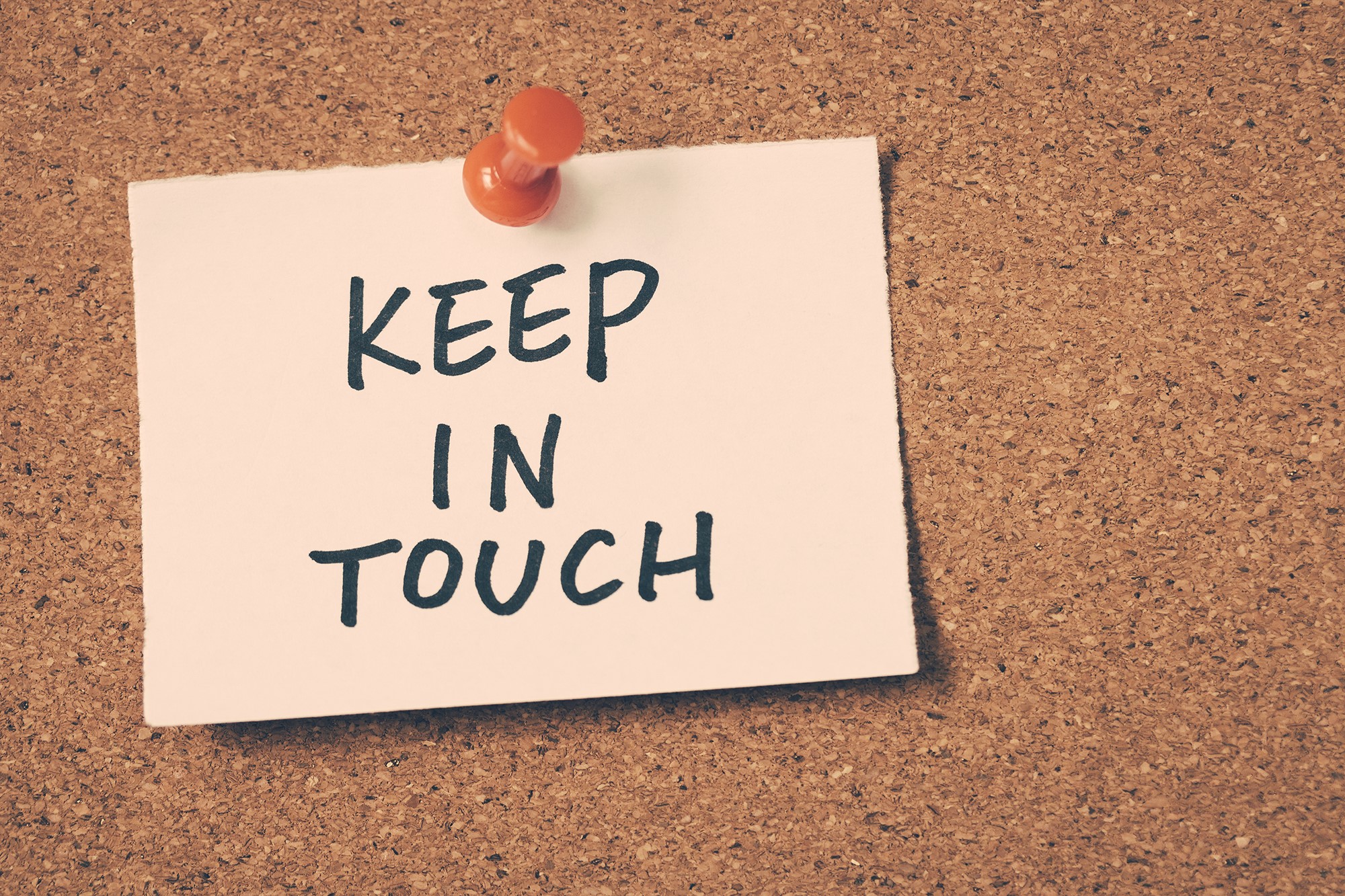 A photo of a sign that reads "Keep in Touch" pinned to a corkboard