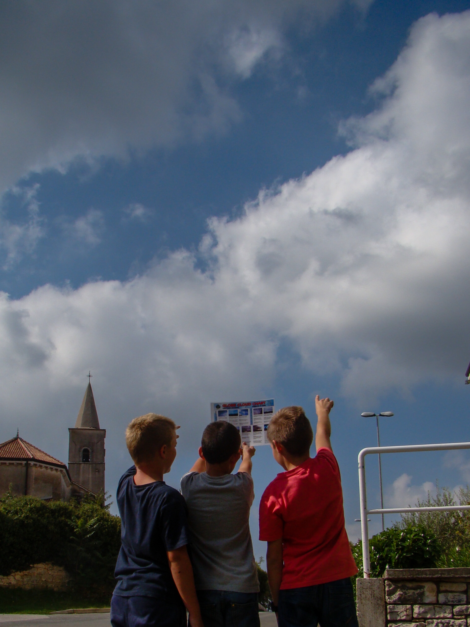 Three young students outside pointing up at clouds while looking at a cloud identification guide.
