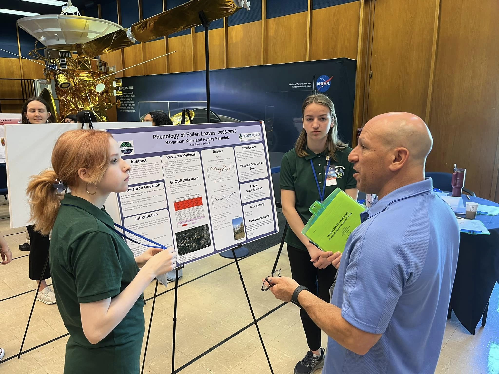 Two students and a reviewer discuss student GLOBE research in front of a poster.