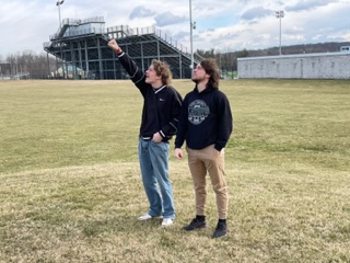 two students stand in a field looking into the sky, one is pointing at the sky; they are collecting cloud data