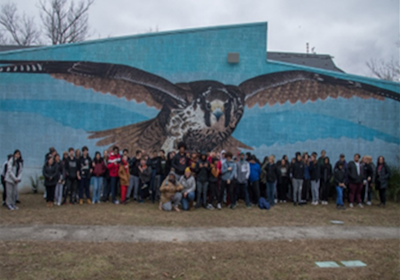 A group of students stands in front of a wall painted with a large bird of prey.