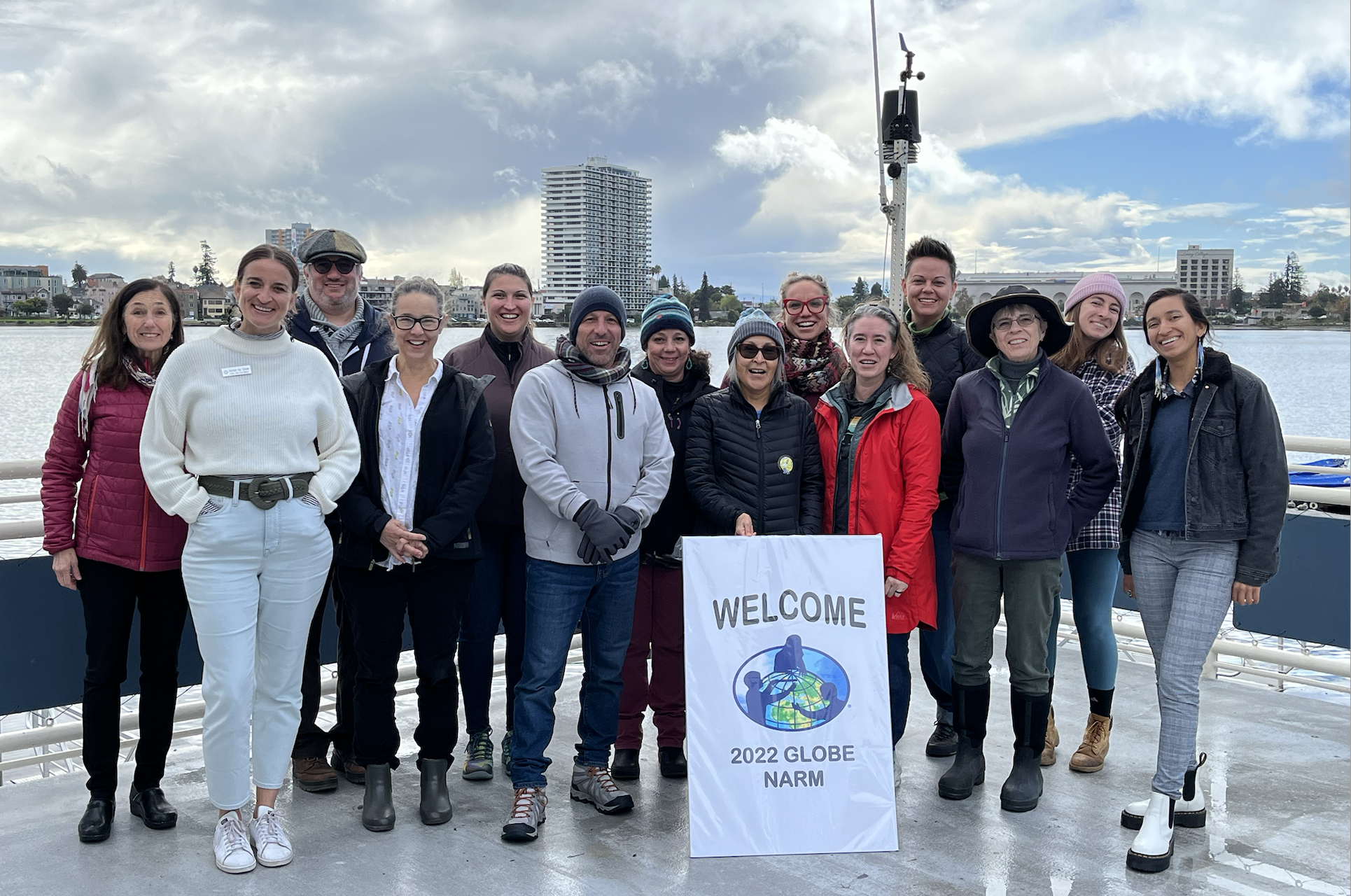 Fourteen participants at the Oakland, CA, in-person North American Regional Meeting stand on a dock in front of Lake Merritt with buildings in the background