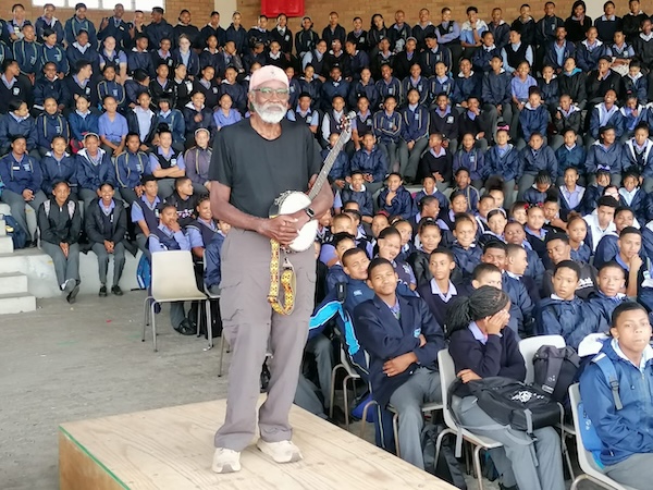 John Francis stands on a platform with his banjo with students in the background.