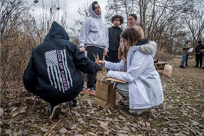 Two students crouch on the ground with leaves while other students stand by watching.