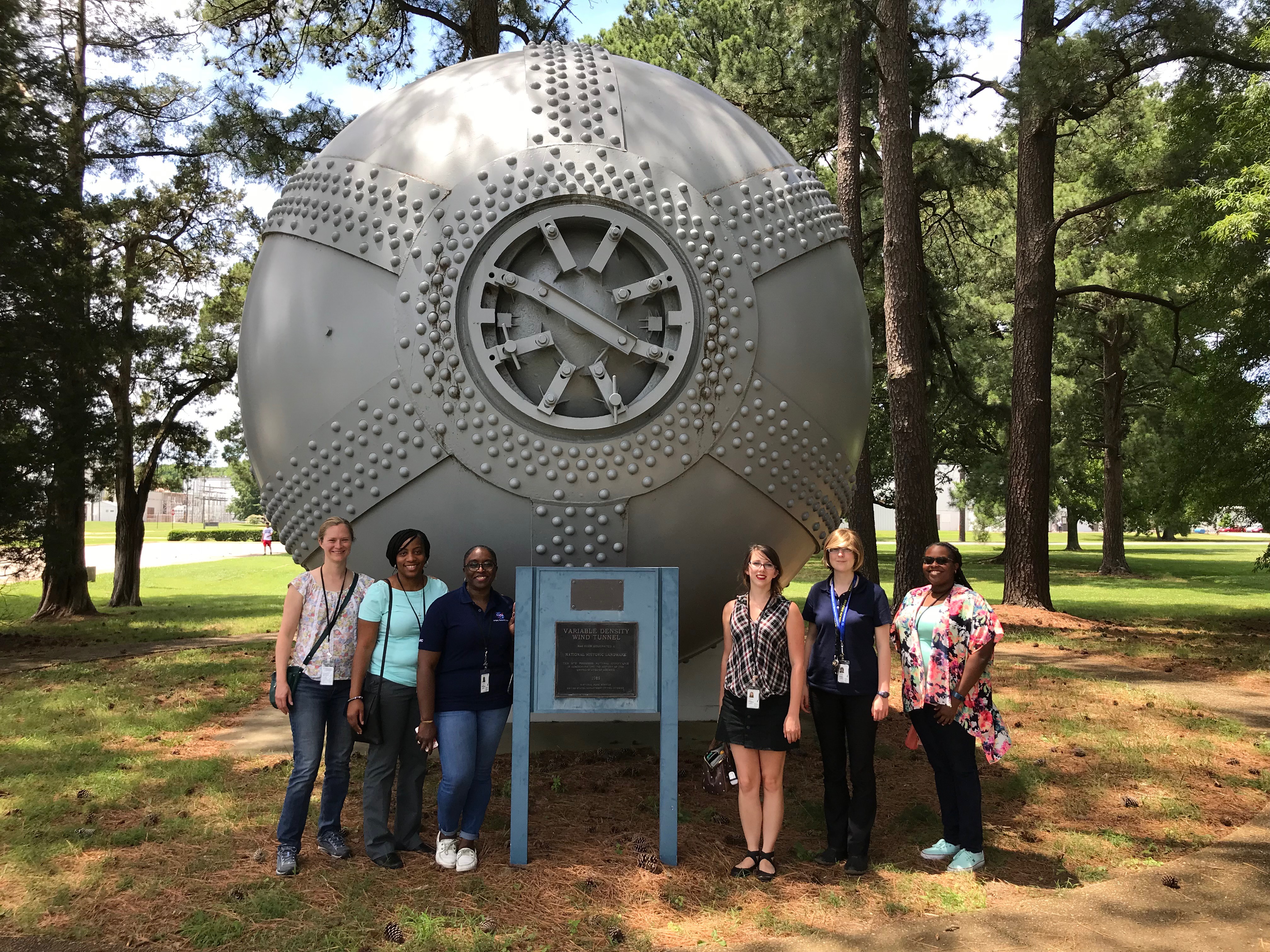 Six female teachers standing in front of a large metal ball - a wind tunnel - outside on a sunny day in a pine woodland.