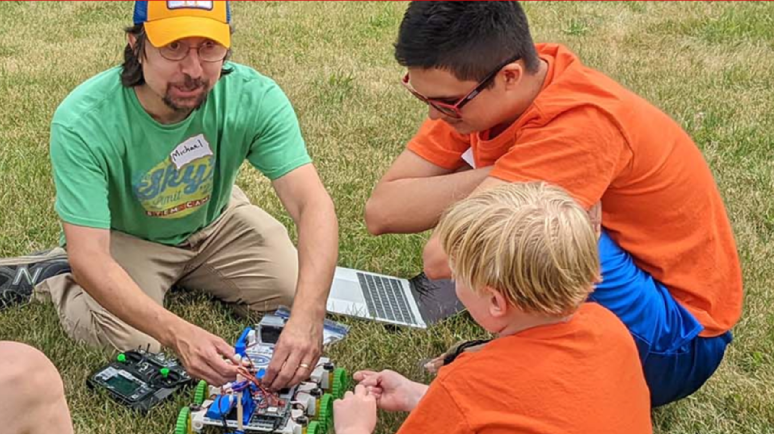 Dr. Michael Notaro sits on the grass with students as he works on a remote-controlled rover that takes GLOBE measurements