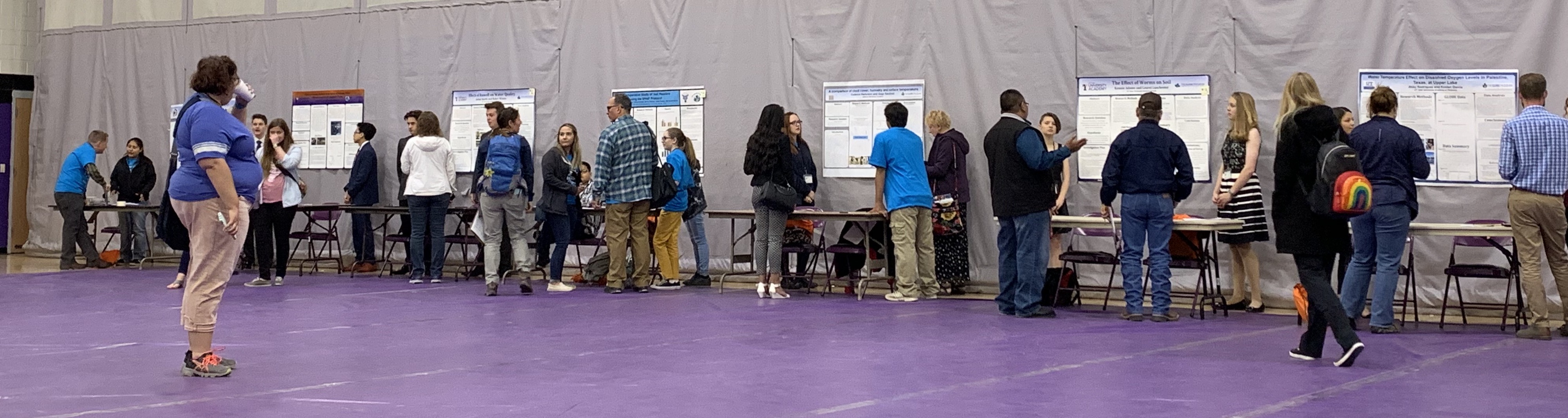 a gym with posters taped to the wall with students presenting to adults and other students along it.