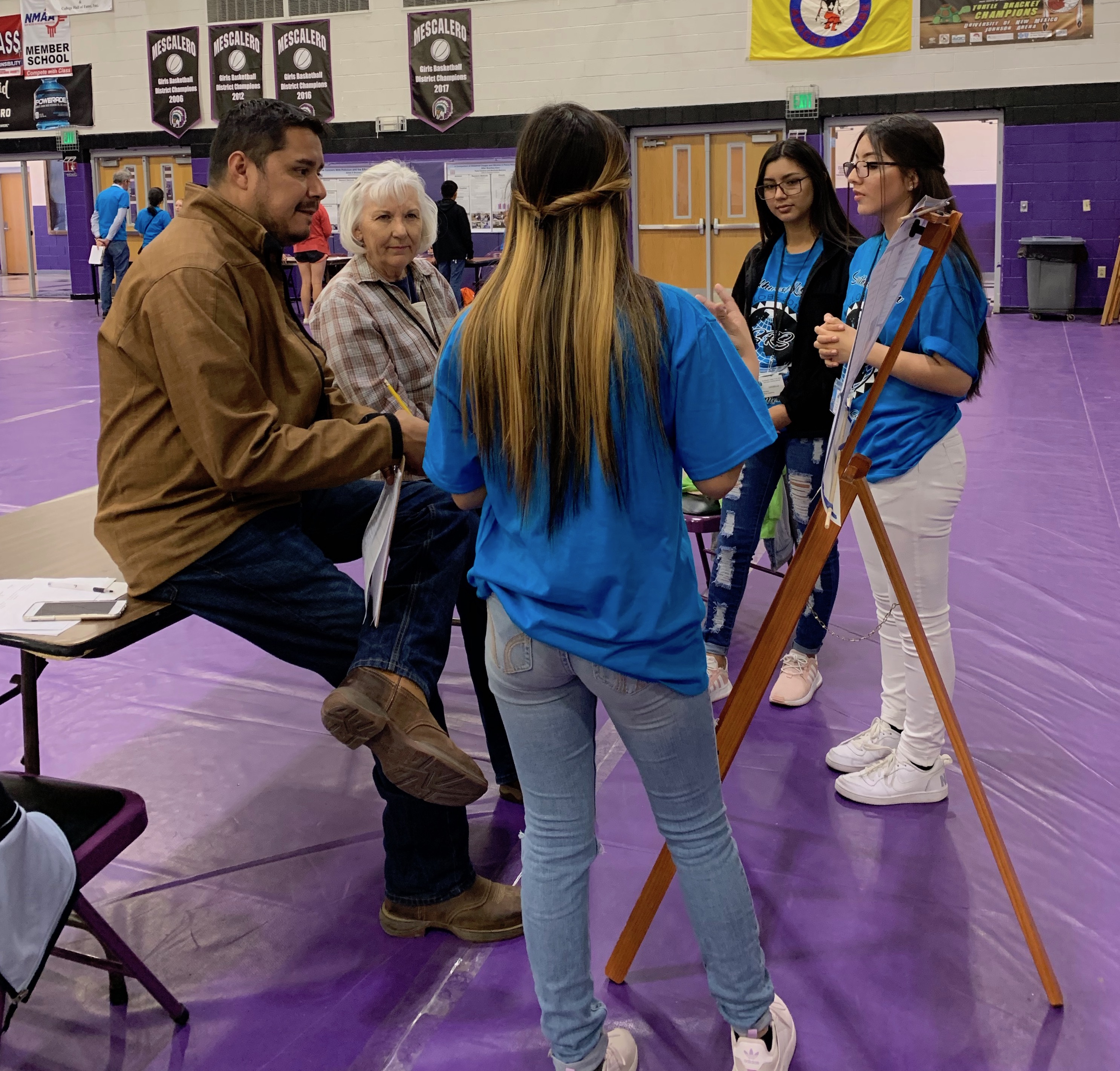 Two reviewers talking to three female students from Oklahoma about their project which is described on a poster and pinned to an easle.