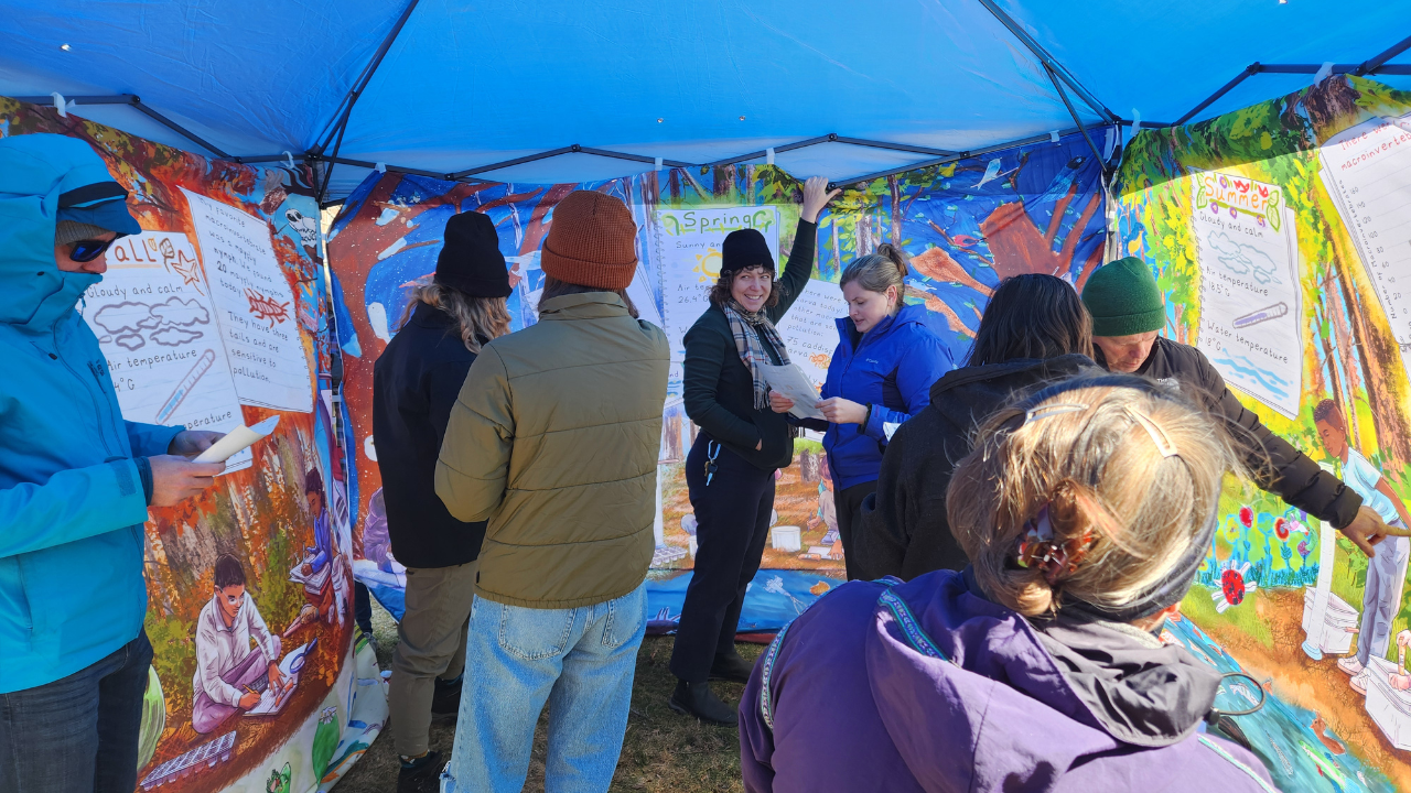 adults stand under a pop-up tent with murals on three sides as they conduct a scavenger hunt