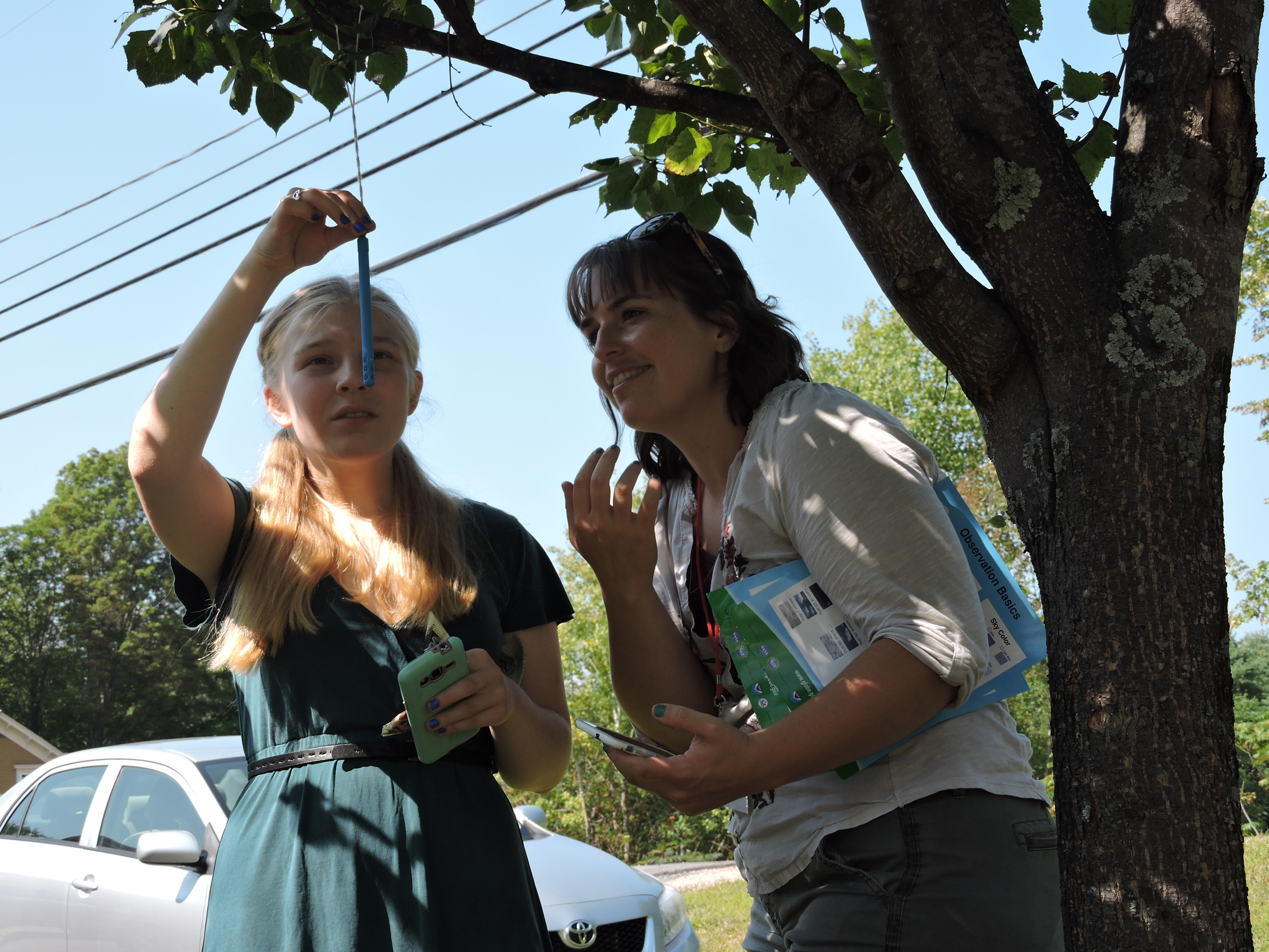 female grad student and young female student reading the thermometer under the shade of a tree