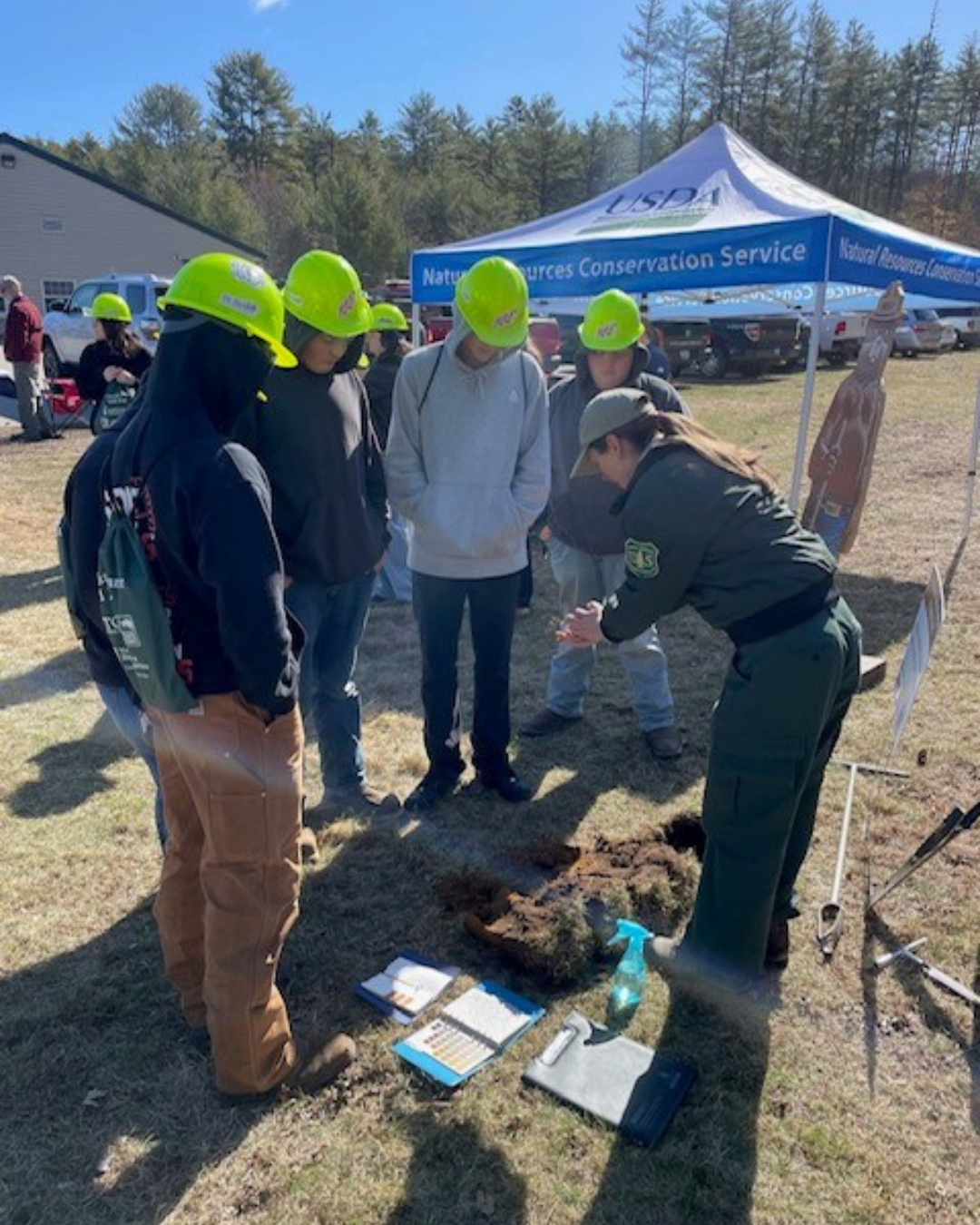 Maddy Czymmek, a soil scientist with the White Mountain National Forest, stands with students at a small soil pit. she has a sample of soil in her hand and color charts, a spray bottle, and other tools on the ground nearby.