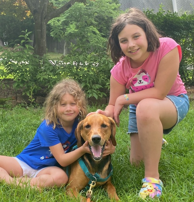 Two girls and a dog on grass, looking at the camera.