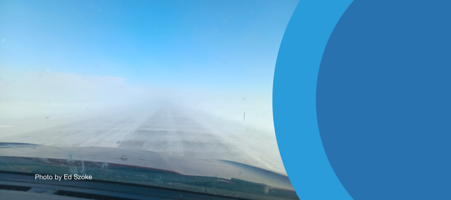 A dash cam image of a road obscured by blowing snow, with blue sky visible above.
