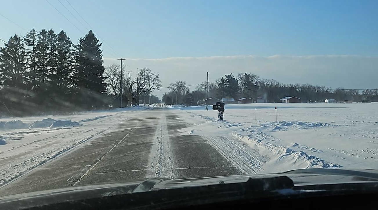 Landscape scene showing snow drifting across a roadway to the left of a snow-covered field.