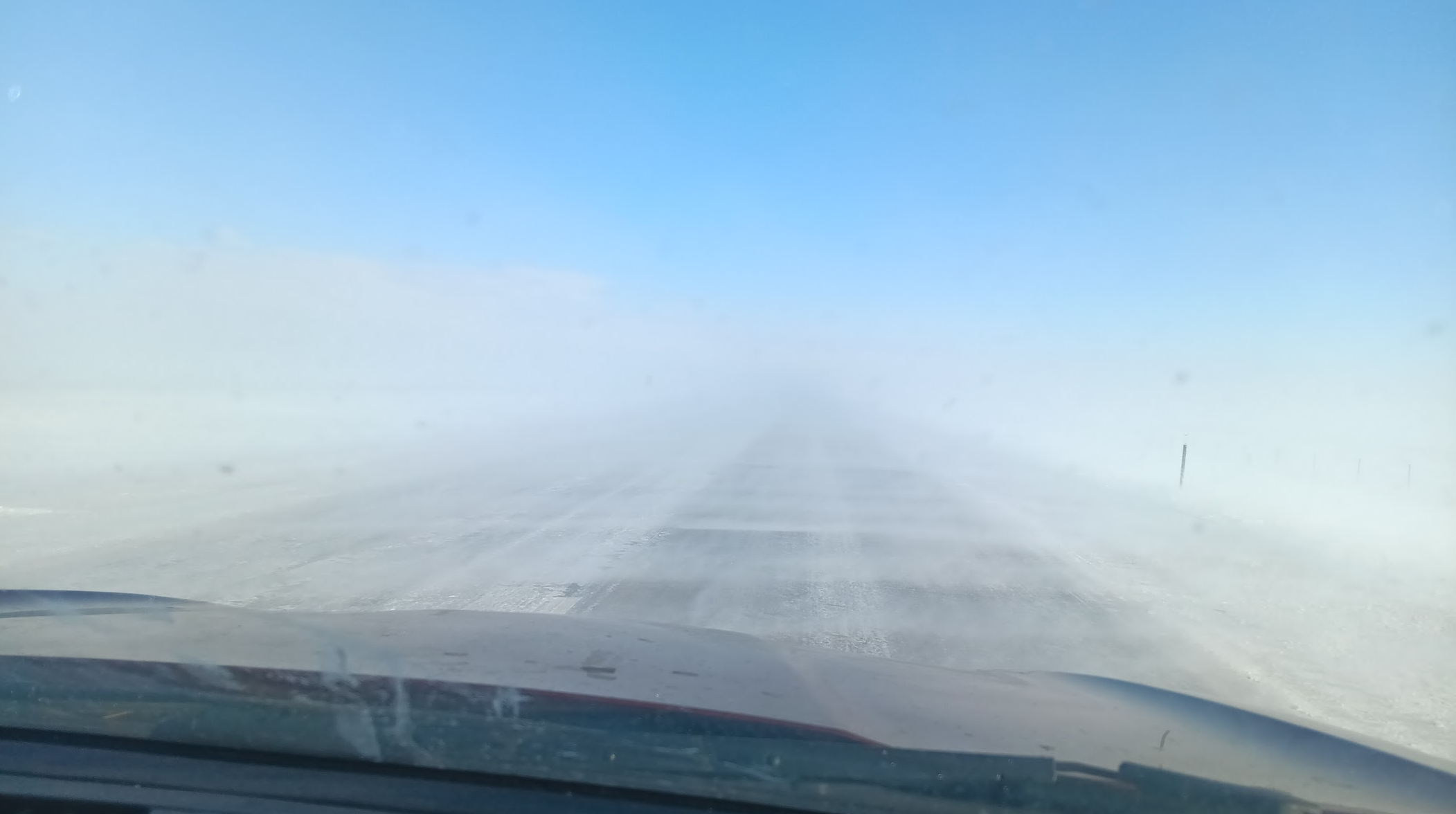 A snowy highway is seen through the windshield of a car. Blowing snow obscures the view in front of the car, but blue sky is visible above.