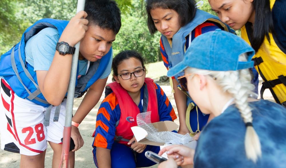 Four students in life jackets watch a teacher explaining a scientific instrument outside.