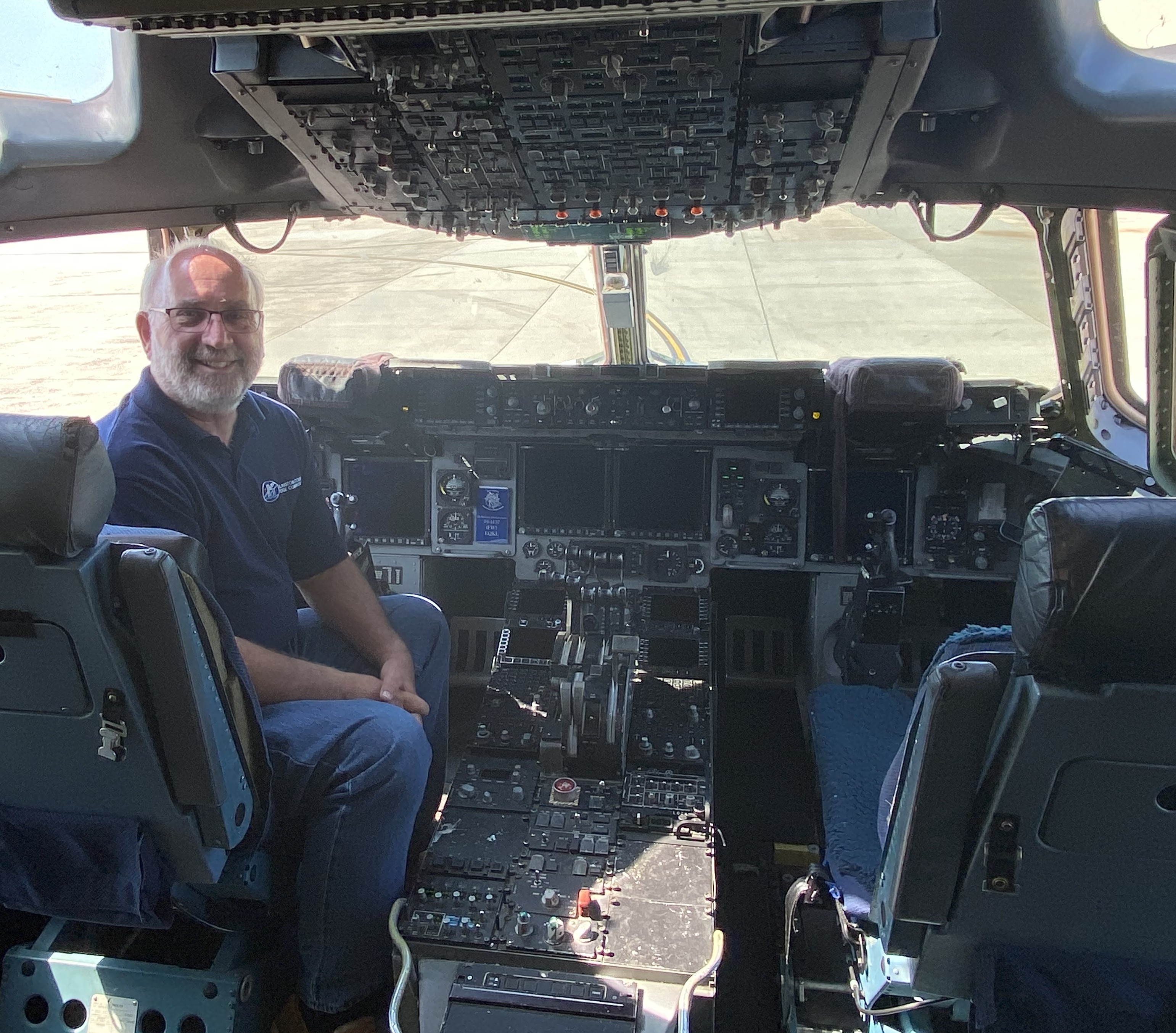 John Moore sits in the cockpit of a C-17; there are many controls in the cockpit; a runway is visible through the cockpit windows