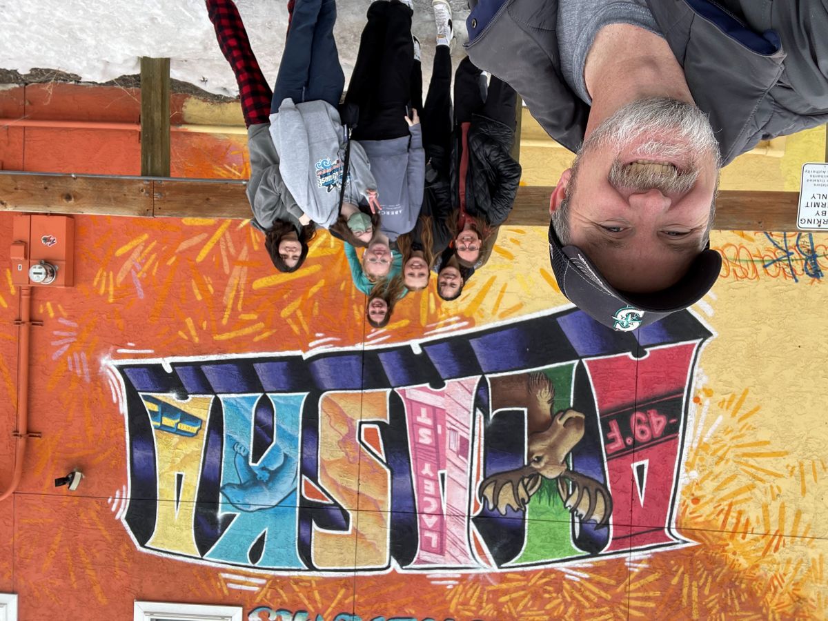 Bill Becker takes a selfie with his students in front of a colorful Alaska mural