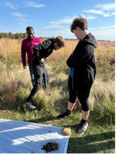 One student digs in the prairie soil while others observe; a sheet on the ground holds soil samples.