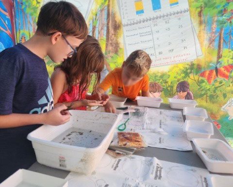 Young campers gather around a table to identify freshwater macroinvertebrates, with the Earth Around Us water tent in the background.