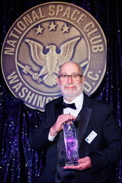 John Moore holds a crystal award while standing in front of a National Space Club placard at the Robert H. Goddard Memorial Dinner on March 13, 2026.