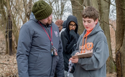 A teacher stands next to a student and looks at their clipboard.