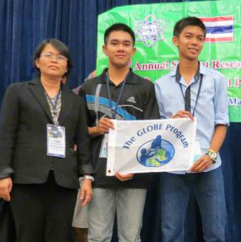 Two students and a teacher are standing in front of a curtain and sign. The students are holding a GLOBE Program flag and smiling.
