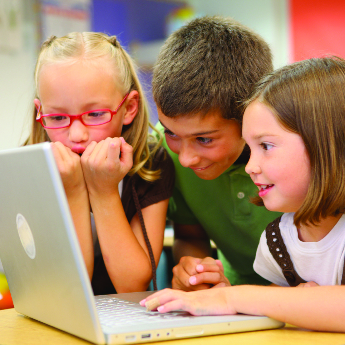 Three children are sitting around a laptop, smiling and looking at the screen. 