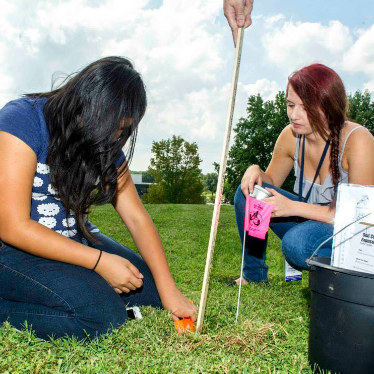 Two women kneel on grass. One woman holds an orange shovel and is digging into soil while the other holds a bag of seeds.