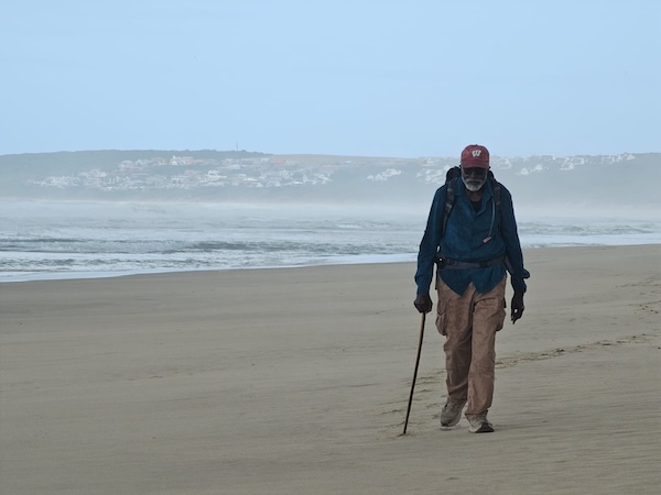 John Francis walking with a cane on a beach with water in the background.