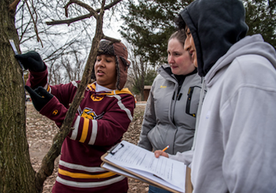 Students standing by a tree, holding a measuring tape and clipboard.